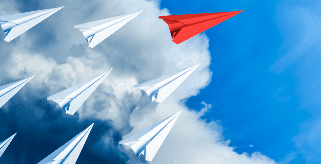 red paper airplane flies ahead of white paper airplanes in the sky