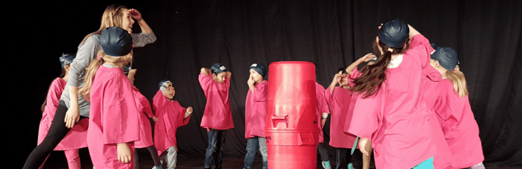 children in pink shirts wander and look at two red garbage bins stacked on top of eachother