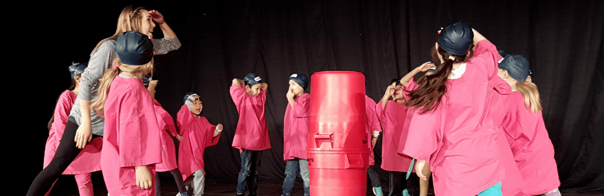 children in pink shirts wander and look at two red garbage bins stacked on top of eachother